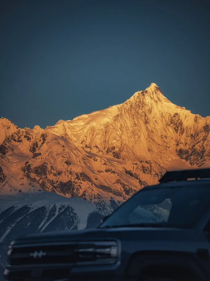 金色光线照亮雪山尖峰,近处汽车映衬深蓝色天空。